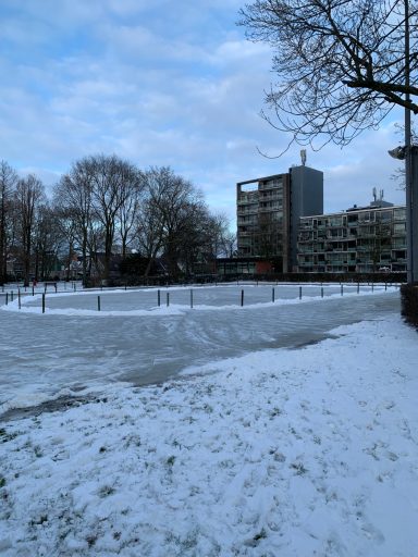 Sneeuwbedekte lege speelplaats met bomen en gebouwen op de achtergrond.